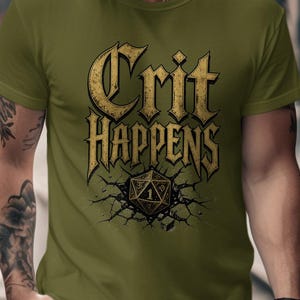 Op de afbeelding: Olijfgroen t-shirt met de tekst "Crit Happens" in een gouden, gotische lettertype. Daaronder een afbeelding van een gebarsten oppervlak met een gouden D20 dobbelsteen. Het t-shirt heeft korte mouwen.