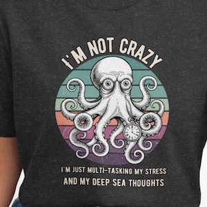 May include: Dark gray t-shirt featuring a white octopus illustration with a vintage sunset background. The text reads "I'M NOT CRAZY" above the octopus and "I'M JUST MULTI-TASKING MY STRESS AND MY DEEP SEA THOUGHTS" below.