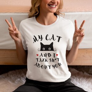 May include: White t-shirt featuring the text "MY CAT AND I TALK SH*T ABOUT YOU" and a black cat graphic. The cat graphic has two eyes and two whiskers. The text is in a black, handwritten style font. Two red hearts are included in the design.