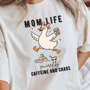 Könnte beinhalten: Cremefarbenes T-Shirt mit der Aufschrift "MOM LIFE" über einer Cartoon-Ente mit rosa Schleife und Kaffeetasse. Die Ente rennt, darunter der Text "powered by CAFFEINE AND CHAOS".