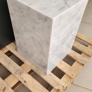 K&ouml;nnte beinhalten: Ein wei&szlig;er Marmor-W&uuml;rfelsockel, der auf einer Holzpalette steht. Der Sockel ist glatt und hat eine hellgraue Maserung.