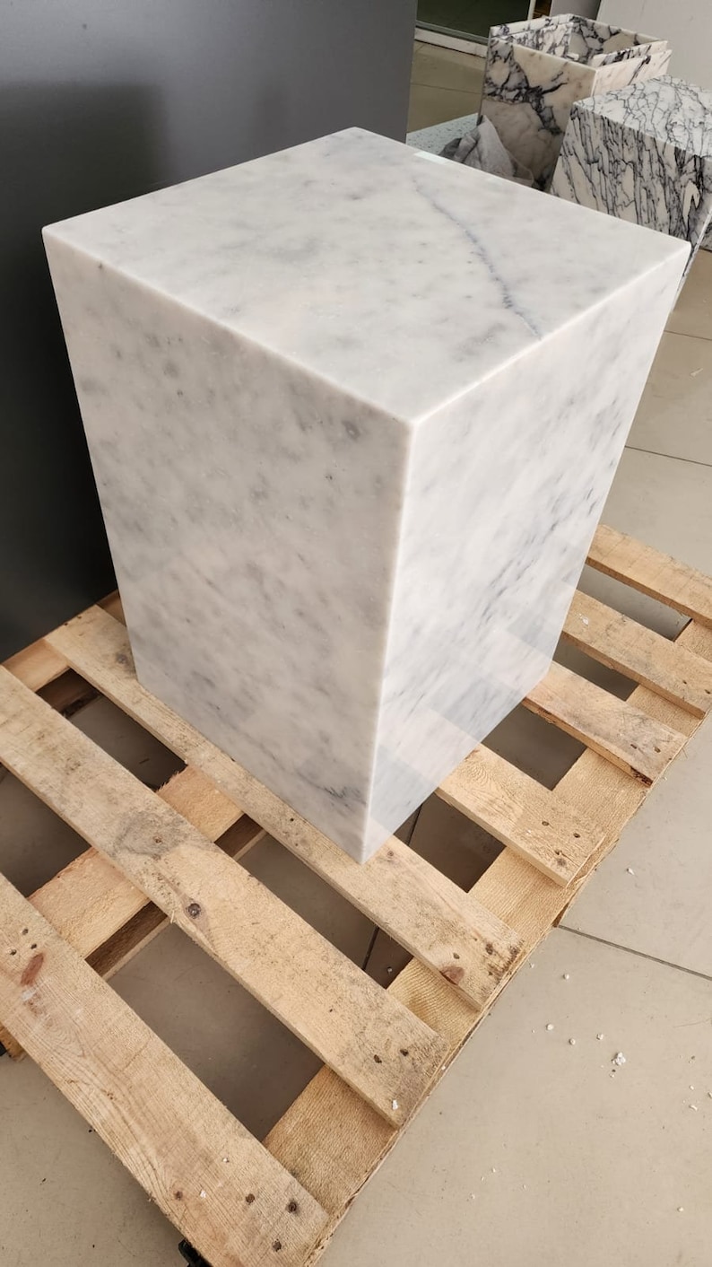 K&ouml;nnte beinhalten: Ein wei&szlig;er Marmor-W&uuml;rfelsockel, der auf einer Holzpalette steht. Der Sockel ist glatt und hat eine hellgraue Maserung.