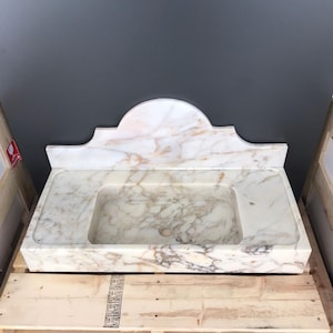 K&ouml;nnte beinhalten: Ein wei&szlig;es Marmor-Waschbecken mit einem dekorativen, gew&ouml;lbten R&uuml;ckspritzschutz. Das Waschbecken steht auf einer Holzpalette.