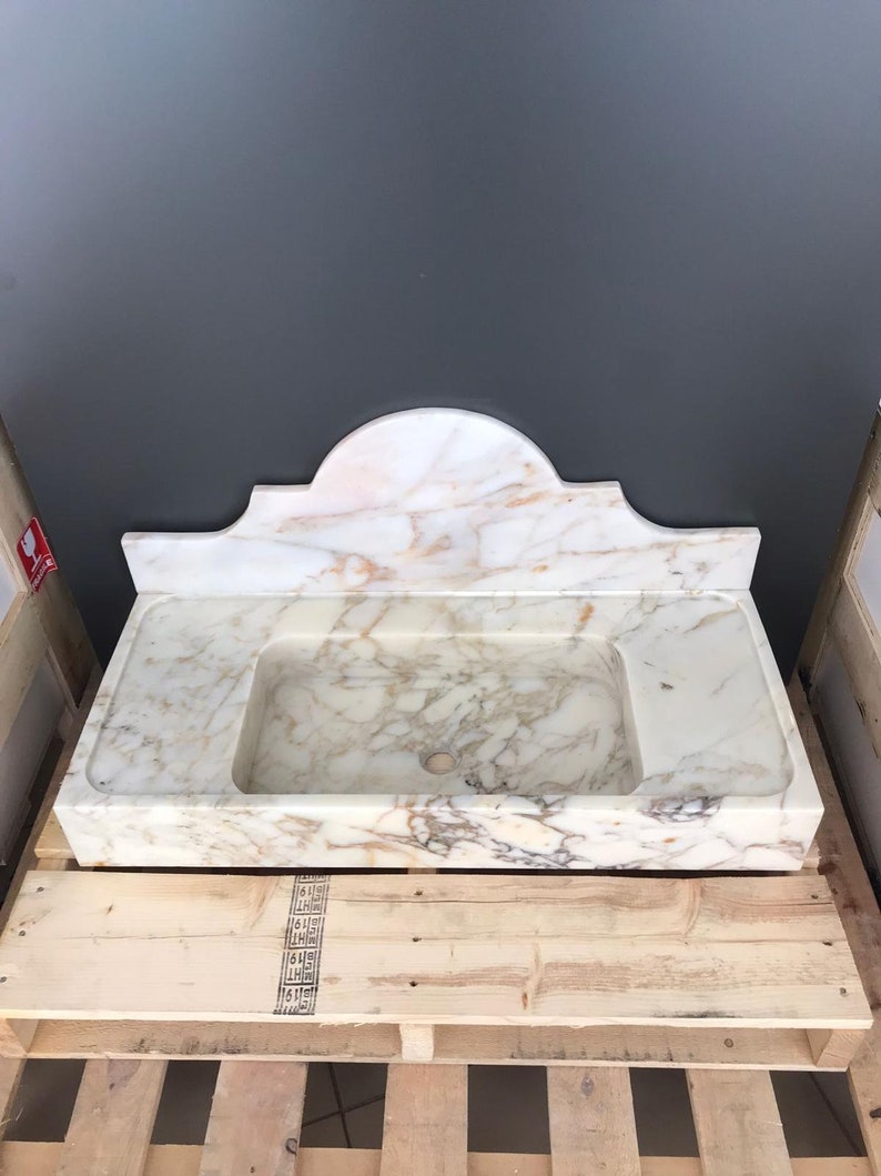 K&ouml;nnte beinhalten: Ein wei&szlig;es Marmor-Waschbecken mit einem dekorativen, gew&ouml;lbten R&uuml;ckspritzschutz. Das Waschbecken steht auf einer Holzpalette.