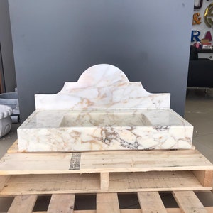 K&ouml;nnte beinhalten: Ein wei&szlig;es Marmor-Waschbecken mit einem dekorativen, gew&ouml;lbten Oberteil. Das Waschbecken steht auf einer Holzpalette.