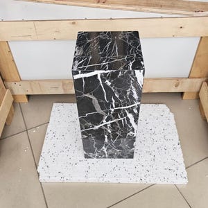 May include: A tall, hexagonal marble pedestal with a black and white veined pattern. The pedestal is set on a white, speckled base. The background includes wooden supports and a white surface.