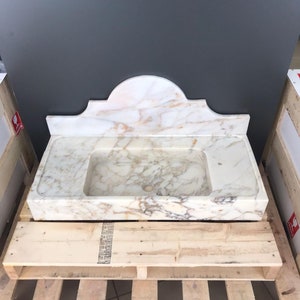 K&ouml;nnte beinhalten: Ein wei&szlig;es Marmor-Waschbecken mit einem dekorativen, gew&ouml;lbten Oberteil. Das Waschbecken steht auf einer Holzpalette.