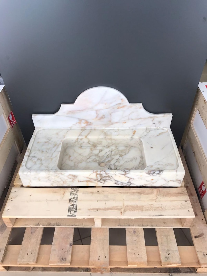K&ouml;nnte beinhalten: Ein wei&szlig;es Marmor-Waschbecken mit einem dekorativen, gew&ouml;lbten Oberteil. Das Waschbecken steht auf einer Holzpalette.