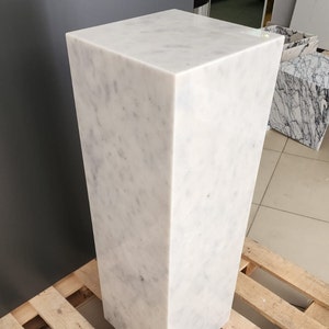K&ouml;nnte beinhalten: Ein wei&szlig;er Marmorsockel mit quadratischer Oberseite und rechteckiger Basis. Der Sockel steht auf einer Holzpalette.