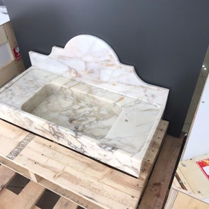 K&ouml;nnte beinhalten: Ein wei&szlig;es Marmor-Waschbecken mit einem dekorativen, gew&ouml;lbten R&uuml;ckspritzschutz. Das Waschbecken steht auf einer Holzpalette.