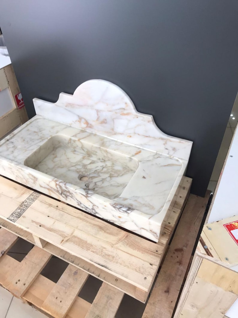 K&ouml;nnte beinhalten: Ein wei&szlig;es Marmor-Waschbecken mit einem dekorativen, gew&ouml;lbten R&uuml;ckspritzschutz. Das Waschbecken steht auf einer Holzpalette.