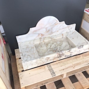 K&ouml;nnte beinhalten: Ein wei&szlig;es Marmor-Waschbecken mit einem dekorativen, gew&ouml;lbten Oberteil. Das Waschbecken steht auf einer Holzpalette.