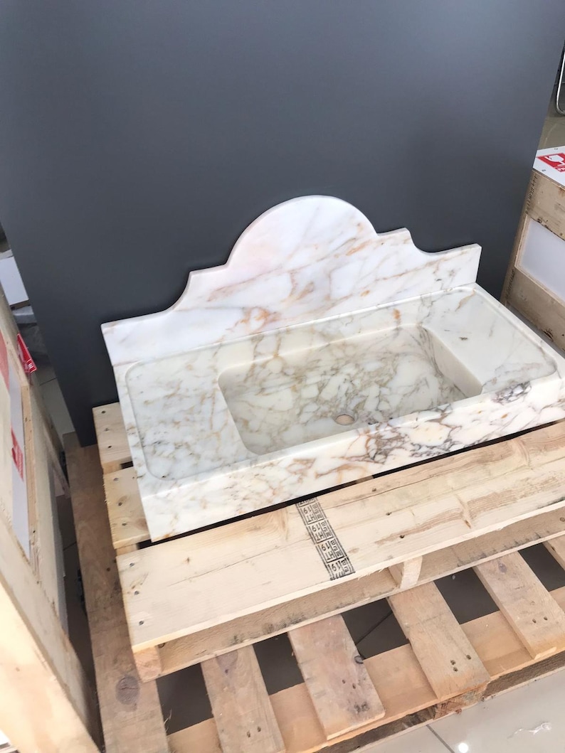 K&ouml;nnte beinhalten: Ein wei&szlig;es Marmor-Waschbecken mit einem dekorativen, gew&ouml;lbten Oberteil. Das Waschbecken steht auf einer Holzpalette.