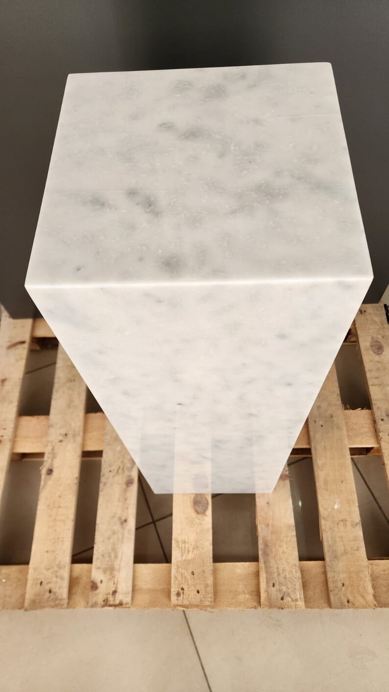 K&ouml;nnte beinhalten: Ein wei&szlig;er Marmorsockel mit einer glatten, polierten Oberfl&auml;che. Der Sockel ist rechteckig und steht auf einer Holzpalette.