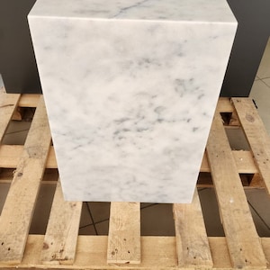 K&ouml;nnte beinhalten: Ein wei&szlig;er Marmor-W&uuml;rfelsockel, der auf einer Holzpalette steht. Der Marmor hat ein hellgraues Adernmuster.
