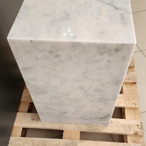 K&ouml;nnte beinhalten: Ein wei&szlig;er Marmor-W&uuml;rfelsockel, der auf einer Holzpalette steht.
