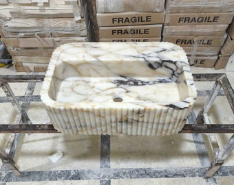 May include: A white marble sink with a rippled edge and a black marble vein running through the centre. The sink is sitting on a metal stand.