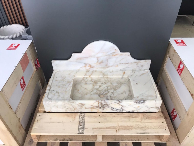 K&ouml;nnte beinhalten: Ein wei&szlig;es Marmor-Waschbecken mit einem dekorativen, gew&ouml;lbten Oberteil. Das Waschbecken steht auf einer Holzpalette.