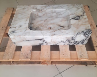 Op de afbeelding: Een witte marmeren wastafel met bruine en grijze aderen. De wastafel is rechthoekig en heeft een afgeronde rand. Hij staat op een houten pallet.