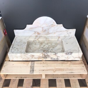 K&ouml;nnte beinhalten: Ein wei&szlig;es Marmor-Waschbecken mit einem dekorativen, gew&ouml;lbten Oberteil. Das Waschbecken steht auf einer Holzpalette.