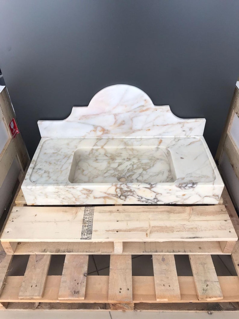 K&ouml;nnte beinhalten: Ein wei&szlig;es Marmor-Waschbecken mit einem dekorativen, gew&ouml;lbten Oberteil. Das Waschbecken steht auf einer Holzpalette.