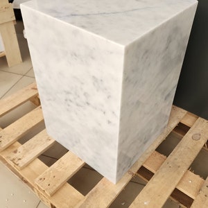 K&ouml;nnte beinhalten: Ein wei&szlig;er Marmor-W&uuml;rfelhocker mit einem hellgrauen Maserungsmuster. Der Hocker steht auf einer Holzpalette.