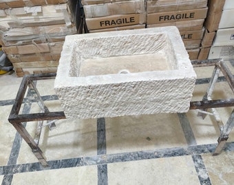May include: A rectangular, light-coloured stone sink with a textured exterior. The sink is supported by a dark metal frame. Cardboard boxes labelled "FRAGILE" are stacked in the background.