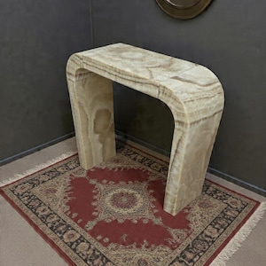 Op de afbeelding: Een console-tafel in watervalstijl gemaakt van gepolijste beige en crèmekleurige onyxsteen. De tafel heeft een glad, doorlopend gebogen ontwerp met zichtbare natuurlijke aders die door het doorschijnende stenen oppervlak lopen.