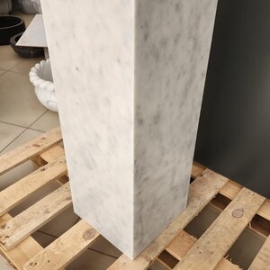 K&ouml;nnte beinhalten: Ein wei&szlig;er Marmorsockel mit quadratischer Oberseite und rechteckiger Basis. Der Sockel steht auf einer Holzpalette.