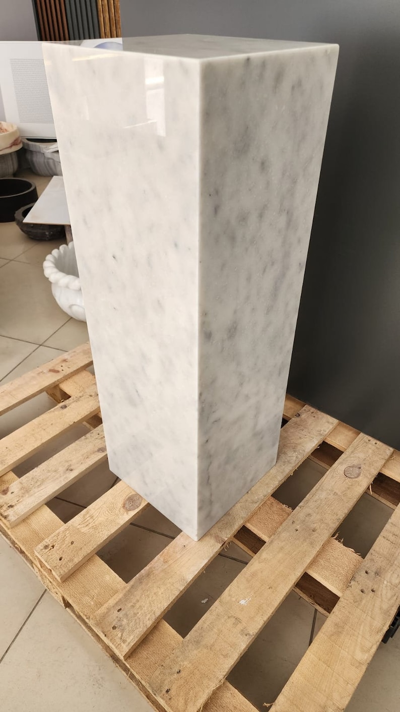 K&ouml;nnte beinhalten: Ein wei&szlig;er Marmorsockel mit quadratischer Oberseite und rechteckiger Basis. Der Sockel steht auf einer Holzpalette.
