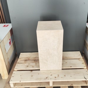 May include: A light beige square pedestal made of textured stone. The pedestal is sitting on a wooden pallet.