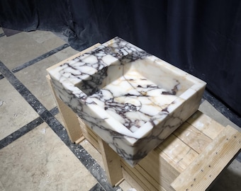 Op de afbeelding: Een witte marmeren wastafel met paarse en bruine aderen, die op een houten kist staat. De wastafel is rechthoekig met een vierkant bassin.
