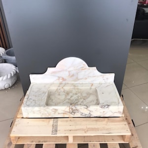 K&ouml;nnte beinhalten: Ein wei&szlig;es Marmor-Waschbecken mit einem dekorativen, gew&ouml;lbten Oberteil. Das Waschbecken steht auf einer Holzpalette.