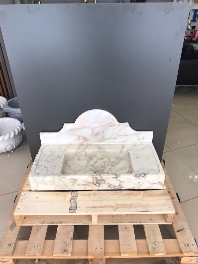 K&ouml;nnte beinhalten: Ein wei&szlig;es Marmor-Waschbecken mit einem dekorativen, gew&ouml;lbten Oberteil. Das Waschbecken steht auf einer Holzpalette.