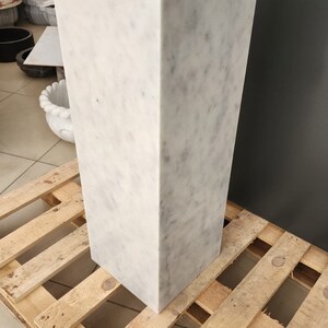 K&ouml;nnte beinhalten: Ein wei&szlig;er Marmorsockel mit quadratischer Oberseite und rechteckiger Basis. Der Sockel steht auf einer Holzpalette.