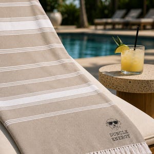May include: Beige and white striped beach towel with a fringe. The towel has a black graphic of a dog wearing sunglasses and the words "DUNCLE ENERGY". A cocktail sits on a side table next to a pool.