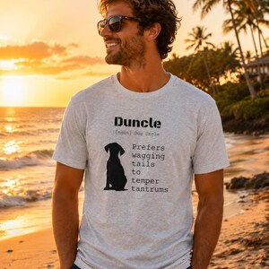May include: Light gray t-shirt with the word "Duncle" and a black silhouette of a dog. The text below reads "Prefers wagging tails to temper tantrums". The shirt is worn by a person on a beach at sunset.