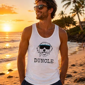 May include: White tank top with a graphic of a dog wearing sunglasses and the word "DUNCLE" in black. The tank top is worn on a beach at sunset.