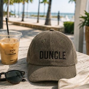 May include: A corduroy baseball cap in olive green with the word "DUNCLE" embroidered in black. The cap sits on a wooden table next to sunglasses, a coffee drink, and a beach towel.