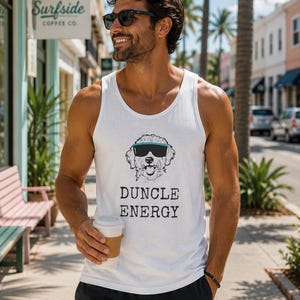 May include: A white tank top with a graphic of a dog wearing sunglasses and the text "DUNCLE ENERGY". The person is wearing black sunglasses and holding a coffee cup. The background includes a coffee shop sign and palm trees.
