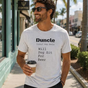 May include: A light gray t-shirt with the word "Duncle" printed in black, defined as "Dog Uncle." Below the definition, the text reads "Will Dog Sit For Beer." The person is holding a coffee cup.