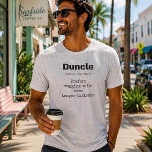 May include: A light gray t-shirt with the word "Duncle" printed in black, along with the definition "Dog Uncle." The shirt also features the phrase "Prefers wagging tails over temper tantrums."
