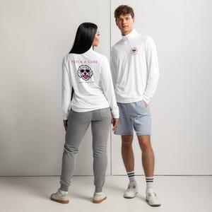 May include: A white long-sleeve quarter-zip pullover featuring a graphic of a dog wearing pink sunglasses on the back with the text FETCH A CURE COASTAL POODLE CLUB and a smaller matching logo on the front chest, paired with gray sweatpants.