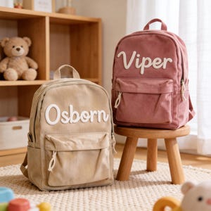 May include: Two corduroy backpacks, one tan and one dusty rose, each with a white embroidered name. The tan backpack says "Osborn" and the dusty rose backpack says "Viper". Both have a front pocket and a top handle.