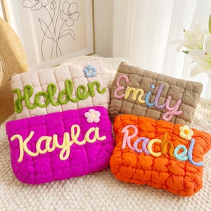 Op de afbeelding: Vier gewatteerde stoffen zakjes in roze, beige en oranje, elk geborduurd met een naam in een contrasterende kleur. De namen zijn "Holder", "Emily", "Kayla" en "Rachel". Elk zakje heeft een klein bloemaccent.