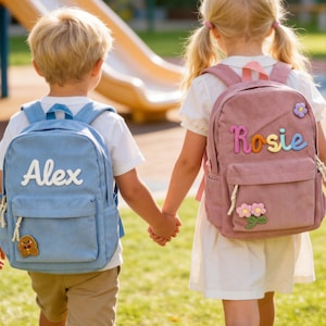 Puede incluir: Dos niños tomados de la mano, cada uno con una mochila de pana. Una es azul claro con el nombre "Alex" en blanco. La otra es rosa con el nombre "Rosie" en letras coloridas. Ambas tienen bolsillos delanteros y parches decorativos.