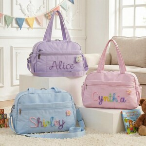 May include: Three pastel-colored baby bags. The bags are lavender, pink, and light blue. Each bag has a name embroidered on the front in a colorful thread. The lavender bag says "Alice", the pink bag says "Cynthia", and the blue bag says "Shirley".