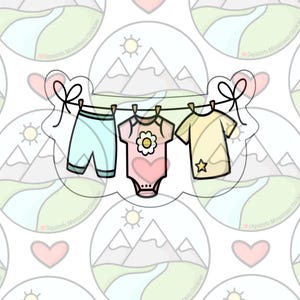 May include: A white background with a line of clothes hanging on a clothesline. The clothes are a light blue pair of shorts, a pink and white onesie with a flower, and a yellow shirt with a star.