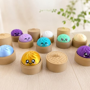 May include: A selection of colourful, smiling dumpling toys in shades of blue, purple, gold, and green, each in a brown steamer basket. The toys are arranged on a light wooden surface.