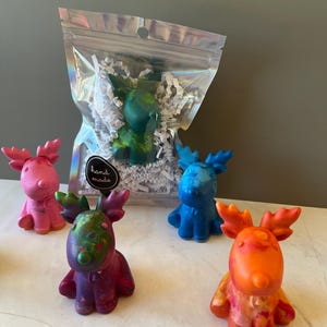 May include: Five colorful reindeer-shaped crayons in a clear plastic bag with white shredded paper. The crayons are pink, blue, green, purple, and orange. The bag has a label that says "hand made".